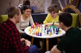 Four people are gathered around a small round table, engaged in a game of chess using brightly colored pieces. The setting appears relaxed and informal, with two men and two women participating. The individuals are focused and appear to be enjoying each other's company in a comfortable indoor environment.