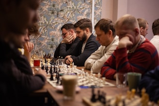 A group of men are deeply focused on playing chess at a table lined with multiple chess boards. The atmosphere is intense as they concentrate on their moves. The room has a patterned wallpaper in the background, and there are various drinks placed on the table accompanying the players.
