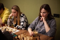 Two women are seated at a table, deeply focused on a game of chess. The woman on the left is wearing a patterned sweater and has her hand resting on her chin. The woman on the right is wearing a sweatshirt and holding a cup of coffee or tea while pondering her next move. There is also a man partly visible on the left wearing a yellow hoodie. Chess pieces are arranged on the boards in front of them, suggesting an intense and strategic game. There is also a smartphone on the table.