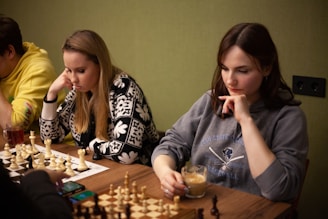 Two women are seated at a table, deeply focused on a game of chess. The woman on the left is wearing a patterned sweater and has her hand resting on her chin. The woman on the right is wearing a sweatshirt and holding a cup of coffee or tea while pondering her next move. There is also a man partly visible on the left wearing a yellow hoodie. Chess pieces are arranged on the boards in front of them, suggesting an intense and strategic game. There is also a smartphone on the table.