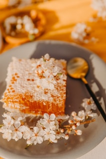 A piece of honeycomb on a gray plate, decorated with white blooming flowers. A gold and black spoon is placed next to the honeycomb. The background is softly blurred with a warm yellow hue.