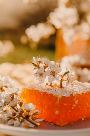 A close-up view of a piece of honeycomb surrounded by delicate white flowers, bathed in warm, golden sunlight. The honeycomb appears richly golden and translucent, while the flowers have small, intricate details, adding to the natural beauty of the scene.