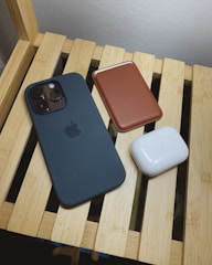 A black smartphone with a camera cluster, a brown leather wallet, and white wireless earbuds are placed on a light wooden slatted surface.
