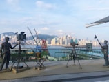 A scenic urban waterfront featuring a row of statues depicting film equipment and crew, including a camera, spotlight, and director's chair. The backdrop is a skyline of skyscrapers and distant mountains under a partly cloudy sky.