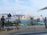 A scenic urban waterfront featuring a row of statues depicting film equipment and crew, including a camera, spotlight, and director's chair. The backdrop is a skyline of skyscrapers and distant mountains under a partly cloudy sky.