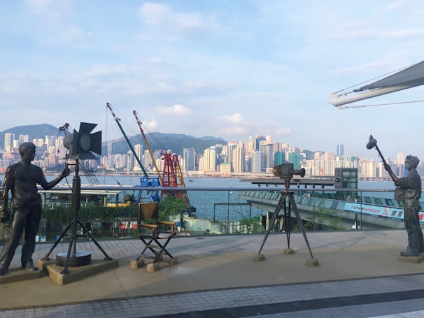 A scenic urban waterfront featuring a row of statues depicting film equipment and crew, including a camera, spotlight, and director's chair. The backdrop is a skyline of skyscrapers and distant mountains under a partly cloudy sky.
