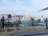 A scenic urban waterfront featuring a row of statues depicting film equipment and crew, including a camera, spotlight, and director's chair. The backdrop is a skyline of skyscrapers and distant mountains under a partly cloudy sky.