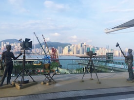 A scenic urban waterfront featuring a row of statues depicting film equipment and crew, including a camera, spotlight, and director's chair. The backdrop is a skyline of skyscrapers and distant mountains under a partly cloudy sky.