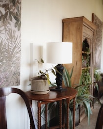 A cozy interior scene features a wooden side table adorned with a potted plant and a decorative lamp with a white shade. The background includes a wooden cabinet with intricate carvings, partially covered by lush green plants. There's also a detailed botanical wall art and a hint of an elegant wooden chair.
