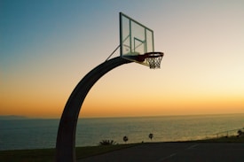 A basketball hoop stands silhouetted against a vibrant sunset. The sky transitions from deep orange to pale yellow as it meets the calm ocean at the horizon. Palm trees dot the landscape, adding to the scenic coastal view.