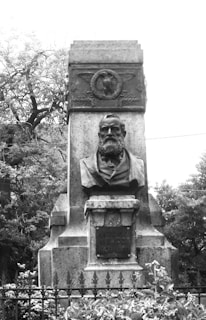 A black and white photograph of a stone monument featuring a bust of an elderly man with a beard. The monument includes an inscription with the name 'Antonio Meucci' and the years '1808-1889'. Surrounding the monument are trees and foliage.