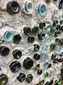 A wall constructed with multiple glass bottle bottoms of various colors, including green, brown, and clear, embedded into a textured cement surface. The glass creates a pattern with light reflecting off the curved surfaces.