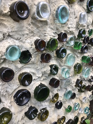 A wall constructed with multiple glass bottle bottoms of various colors, including green, brown, and clear, embedded into a textured cement surface. The glass creates a pattern with light reflecting off the curved surfaces.