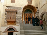 A group of people are gathered on the steps leading up to a historic building with prominent architectural features, including a large wooden door with decorative patterns and stone walls. There is an ornate wooden window on the upper level and a sign for 'St. George Shrine' under a brick archway.