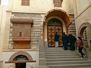 A group of people are gathered on the steps leading up to a historic building with prominent architectural features, including a large wooden door with decorative patterns and stone walls. There is an ornate wooden window on the upper level and a sign for 'St. George Shrine' under a brick archway.