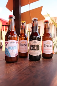 Four glass bottles of craft beer with different labels are displayed on a wooden table. The background shows an outdoor setting with red umbrellas, plants, and rustic buildings.
