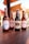 Four glass bottles of craft beer with different labels are displayed on a wooden table. The background shows an outdoor setting with red umbrellas, plants, and rustic buildings.