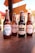 Four glass bottles of craft beer with different labels are displayed on a wooden table. The background shows an outdoor setting with red umbrellas, plants, and rustic buildings.