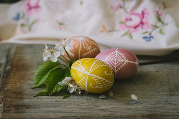 Three intricately decorated Easter eggs are placed on a rustic wooden surface. The eggs feature delicate white patterns on yellow, pink, and orange backgrounds. Next to the eggs, there are fresh green leaves and small white blossoms. A fabric with floral embroidery is softly draped in the background.