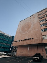 A large building with a modern architectural design features a circular artistic pattern on its facade. Below the pattern, text with Cyrillic characters is displayed. The building is situated next to another structure with a glass exterior, and several cars are parked in a lot nearby. The sky is clear, indicating daytime.
