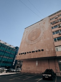 A large building with a modern architectural design features a circular artistic pattern on its facade. Below the pattern, text with Cyrillic characters is displayed. The building is situated next to another structure with a glass exterior, and several cars are parked in a lot nearby. The sky is clear, indicating daytime.