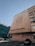 A large building with a modern architectural design features a circular artistic pattern on its facade. Below the pattern, text with Cyrillic characters is displayed. The building is situated next to another structure with a glass exterior, and several cars are parked in a lot nearby. The sky is clear, indicating daytime.