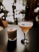Two elegant cocktails are placed on a dark surface with a bokeh effect of warm, soft lighting in the background. The drink on the left is in a short glass with a frothy top and colorful sprinkles. The drink on the right is in a tall, stemmed glass with a smooth, creamy appearance and a decorative item resembling a star anise.