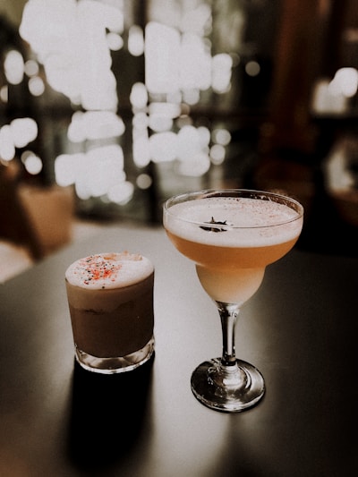 Two elegant cocktails are placed on a dark surface with a bokeh effect of warm, soft lighting in the background. The drink on the left is in a short glass with a frothy top and colorful sprinkles. The drink on the right is in a tall, stemmed glass with a smooth, creamy appearance and a decorative item resembling a star anise.
