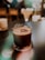 A glass filled with a dark beverage, garnished with a dried citrus slice and sprigs of herbs, sits on a reflective surface. The background is softly blurred with warm light bokeh, creating a cozy atmosphere.