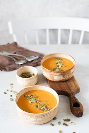 Two bowls of creamy soup are garnished with green sprouts and pumpkin seeds, placed on a wooden cutting board. Nearby, there is a small bowl filled with additional pumpkin seeds. The setup includes a brown cloth and cutlery in the background, and everything is arranged on a light-colored tabletop, with a white chair faintly visible in the background.