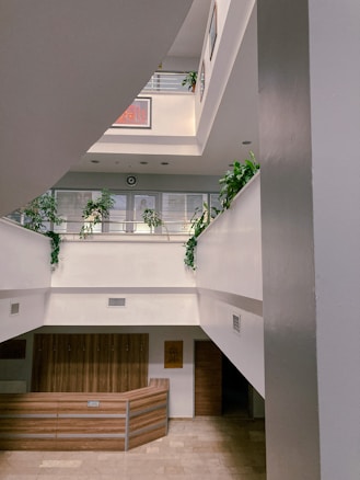A multi-level indoor space with clean architectural lines and abundant natural light. The interior features white walls, wooden accents, and several green plants placed along the upper ledges. A reception desk occupies the ground floor area, and the corridors on each level are open with railings.