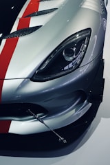 A close-up view of the front section of a sports car featuring a sleek design. The image highlights the headlight and the polished silver color with red and black racing stripes on the car's hood. The car's aesthetic includes aerodynamic vents and a metallic appendage, possibly a support rod or an aerodynamic component.