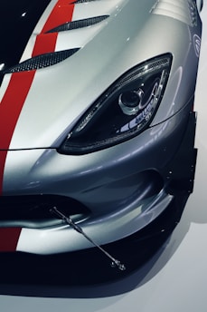 A close-up view of the front section of a sports car featuring a sleek design. The image highlights the headlight and the polished silver color with red and black racing stripes on the car's hood. The car's aesthetic includes aerodynamic vents and a metallic appendage, possibly a support rod or an aerodynamic component.