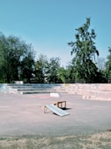 An urban skate park featuring a concrete area with a small wooden ramp in the center. The background contains tiered seating covered in various graffiti, surrounded by lush green trees under a clear blue sky.