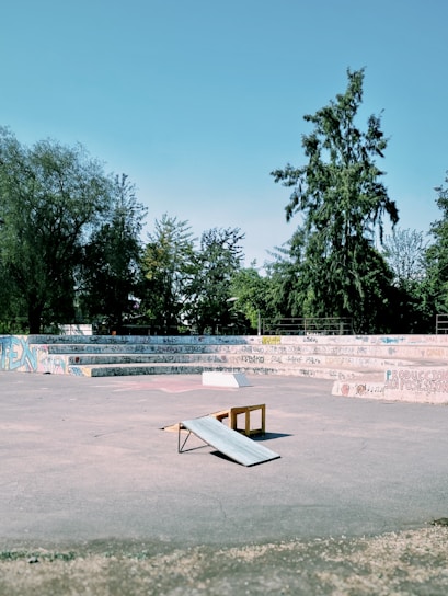 An urban skate park featuring a concrete area with a small wooden ramp in the center. The background contains tiered seating covered in various graffiti, surrounded by lush green trees under a clear blue sky.