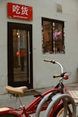 A red bicycle with beige handlebars is parked in front of a small shop. The shop has a red sign with white text above the entrance and a window adorned with stickers. Inside, a neon noodle bowl sign is visible, adding a splash of color against the muted exterior walls.
