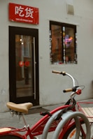 A red bicycle with beige handlebars is parked in front of a small shop. The shop has a red sign with white text above the entrance and a window adorned with stickers. Inside, a neon noodle bowl sign is visible, adding a splash of color against the muted exterior walls.