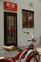 A red bicycle with beige handlebars is parked in front of a small shop. The shop has a red sign with white text above the entrance and a window adorned with stickers. Inside, a neon noodle bowl sign is visible, adding a splash of color against the muted exterior walls.