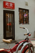 A red bicycle with beige handlebars is parked in front of a small shop. The shop has a red sign with white text above the entrance and a window adorned with stickers. Inside, a neon noodle bowl sign is visible, adding a splash of color against the muted exterior walls.