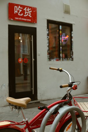 A red bicycle with beige handlebars is parked in front of a small shop. The shop has a red sign with white text above the entrance and a window adorned with stickers. Inside, a neon noodle bowl sign is visible, adding a splash of color against the muted exterior walls.