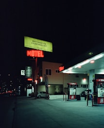 A nighttime street scene featuring a gas station and a motel. Neon signs illuminate the area, including a red 'MOTEL' sign and a green billboard promoting a plant-based diet. The gas station has two pumps and is partially lit by overhead lights. A lone parked car is visible near the motel building.
