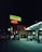A nighttime street scene featuring a gas station and a motel. Neon signs illuminate the area, including a red 'MOTEL' sign and a green billboard promoting a plant-based diet. The gas station has two pumps and is partially lit by overhead lights. A lone parked car is visible near the motel building.
