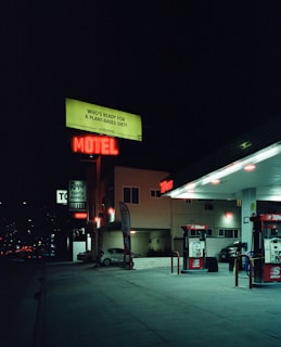 A nighttime street scene featuring a gas station and a motel. Neon signs illuminate the area, including a red 'MOTEL' sign and a green billboard promoting a plant-based diet. The gas station has two pumps and is partially lit by overhead lights. A lone parked car is visible near the motel building.
