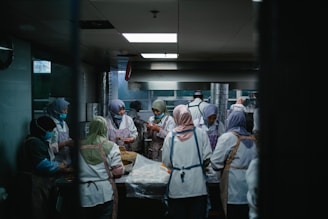 A group of people wearing uniforms and head coverings are working together in a commercial kitchen. They are engaged in preparing food, surrounded by stainless steel appliances and work surfaces. The lighting is dim and gives the room a focused, industrious atmosphere.
