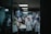 A group of people wearing uniforms and head coverings are working together in a commercial kitchen. They are engaged in preparing food, surrounded by stainless steel appliances and work surfaces. The lighting is dim and gives the room a focused, industrious atmosphere.
