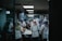 A group of people wearing uniforms and head coverings are working together in a commercial kitchen. They are engaged in preparing food, surrounded by stainless steel appliances and work surfaces. The lighting is dim and gives the room a focused, industrious atmosphere.