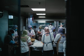 A group of people wearing uniforms and head coverings are working together in a commercial kitchen. They are engaged in preparing food, surrounded by stainless steel appliances and work surfaces. The lighting is dim and gives the room a focused, industrious atmosphere.