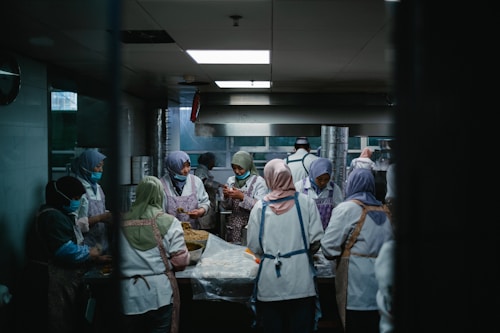 A group of people wearing uniforms and head coverings are working together in a commercial kitchen. They are engaged in preparing food, surrounded by stainless steel appliances and work surfaces. The lighting is dim and gives the room a focused, industrious atmosphere.