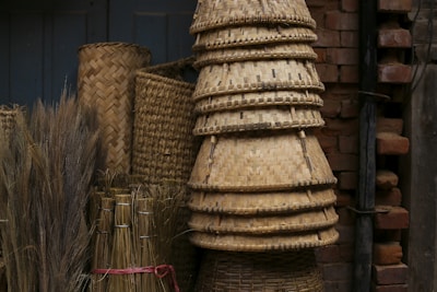 A collection of woven bamboo baskets is stacked against an old brick wall. Various sizes and shapes of baskets are visible, along with bundles of straw or grass tied together. The colors are earthy and the texture of the materials is emphasized.