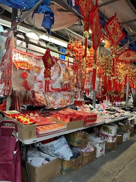 A market stall displaying a variety of red decorative items, likely for a festive occasion. There are ornate hangings featuring intricate designs, shiny gold accents, and assorted lucky symbols. The table is covered with red paper boxes and envelopes, while the floor underneath has cardboard boxes filled with additional goods.